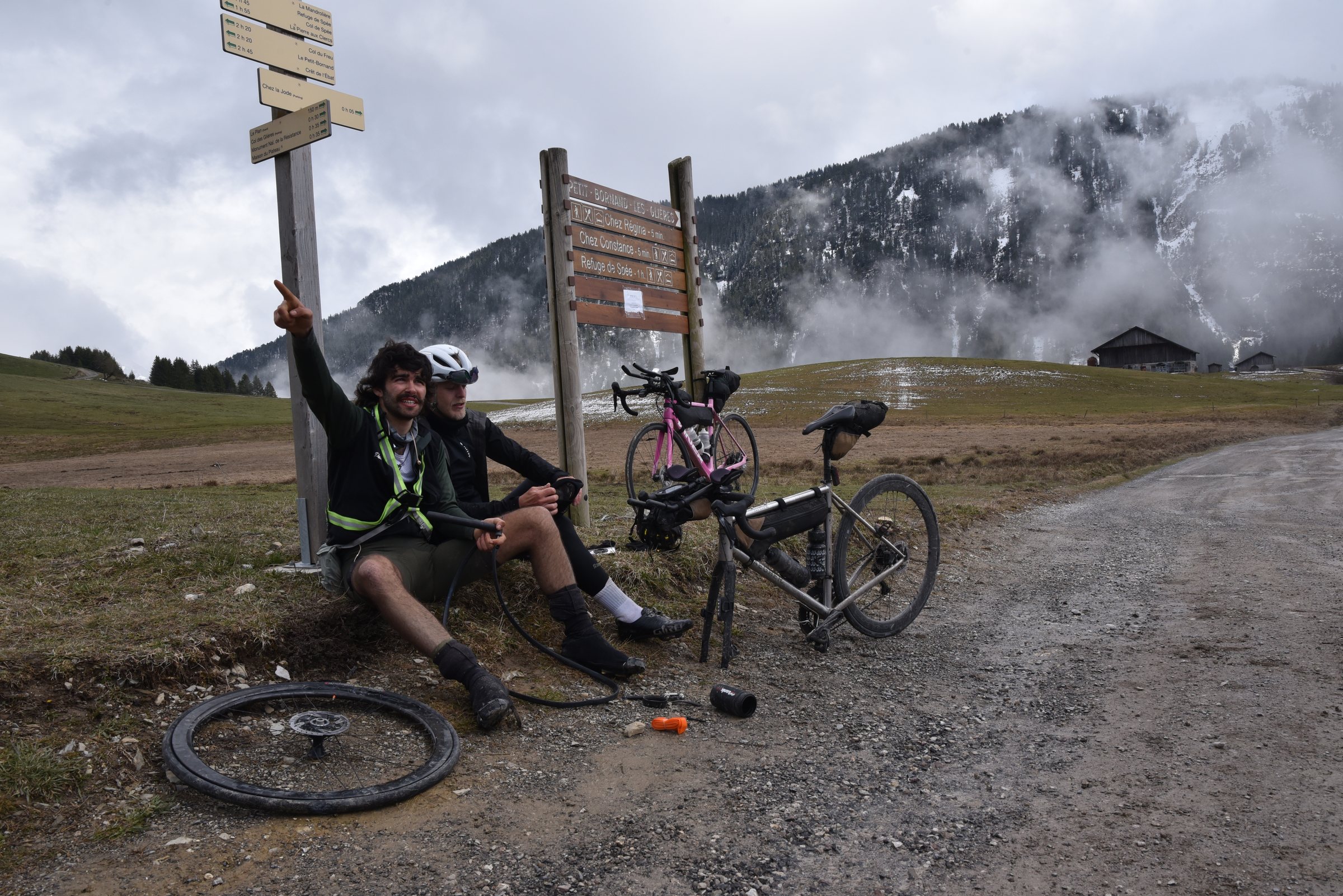 Two riders sitting at a signpost next to a flat tyre, one pointing up, misty snowy mountains behind, during TURN01
