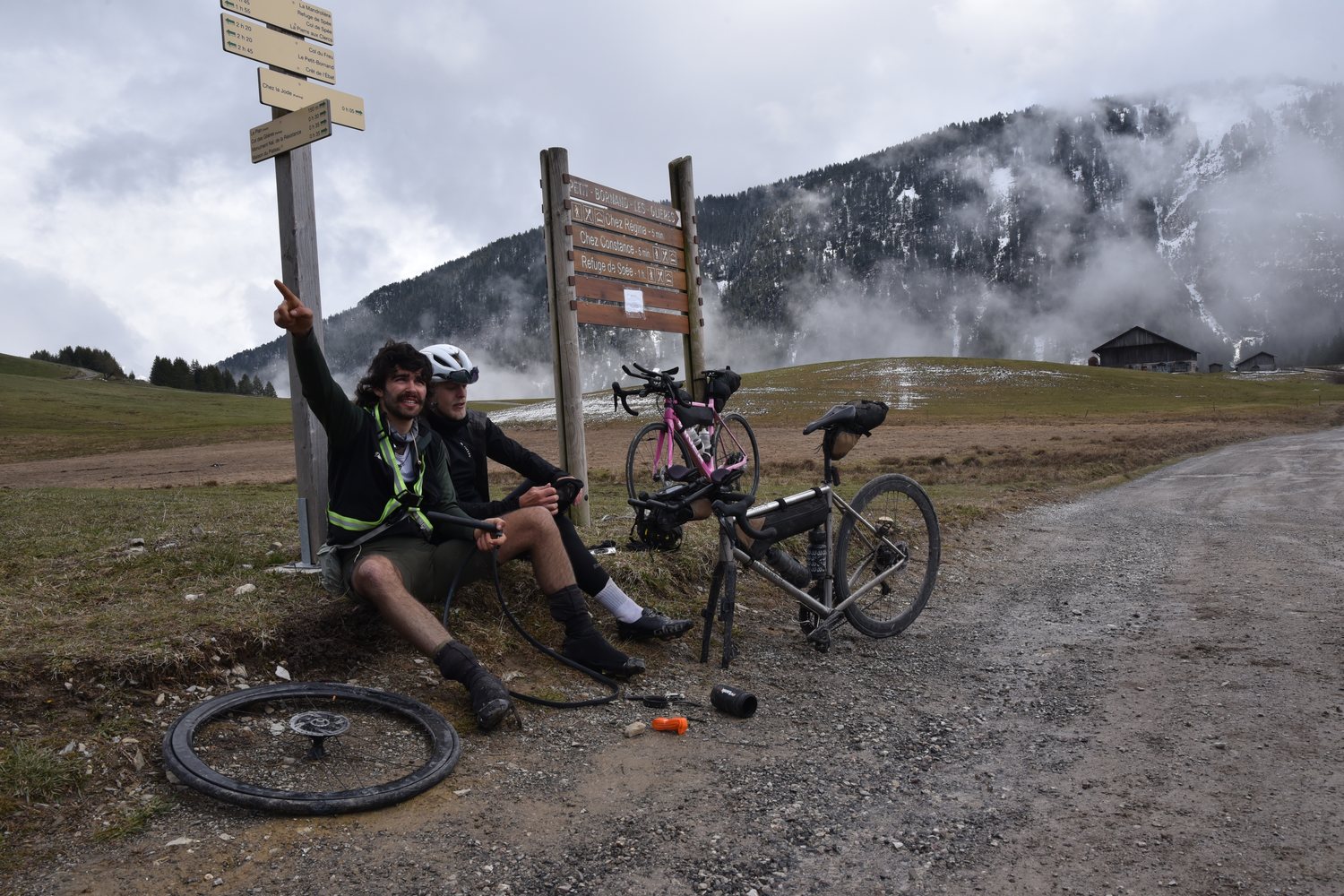 Two riders sitting at a signpost next to a flat tyre, one pointing up, misty snowy mountains behind, during TURN01