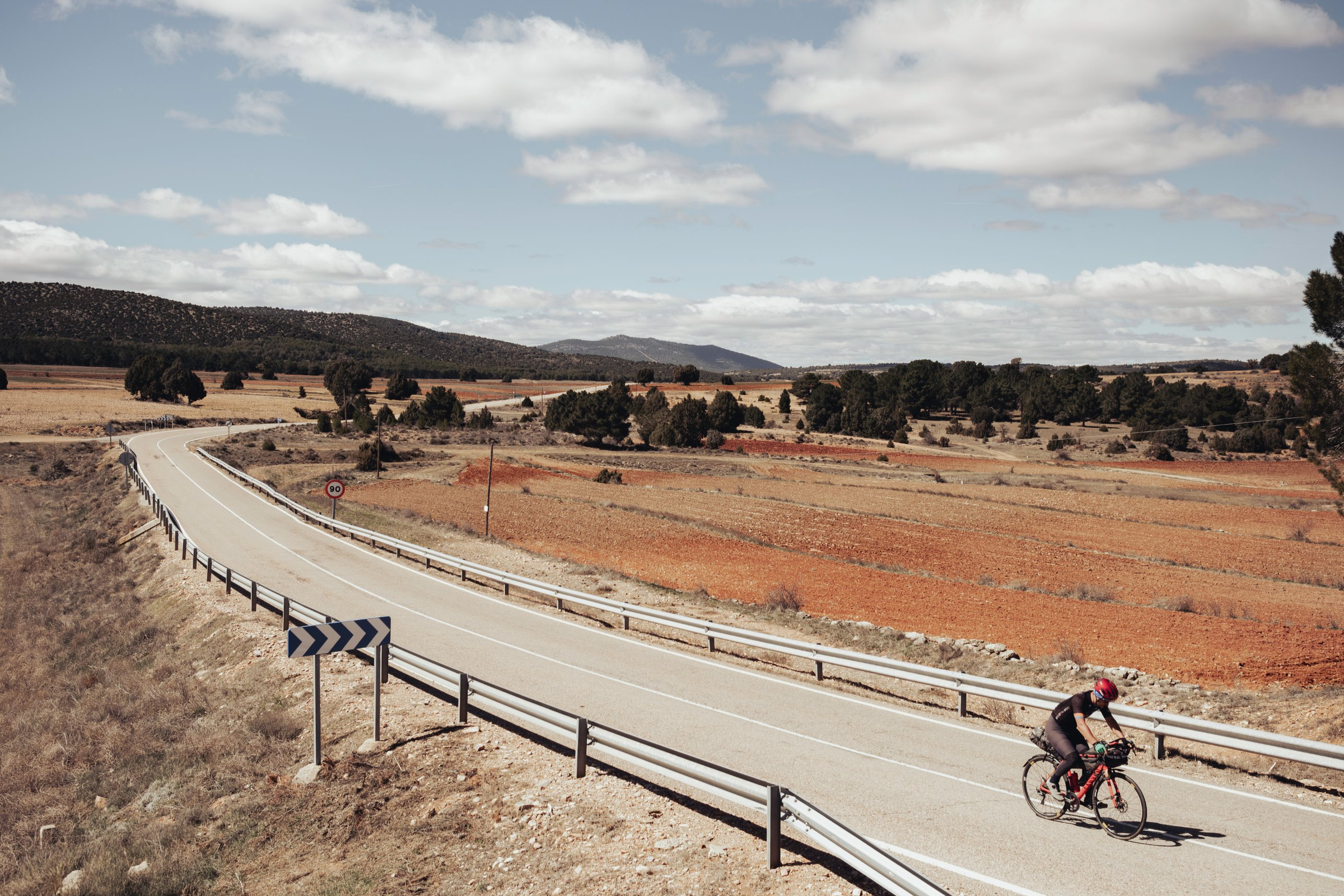 Rider climbing a quiet road through red fields and pine hills